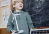 Vertical shot of cheerful primary learner presenting green energy project holding small solar powered wind turbine and big solar panel showing devices to class