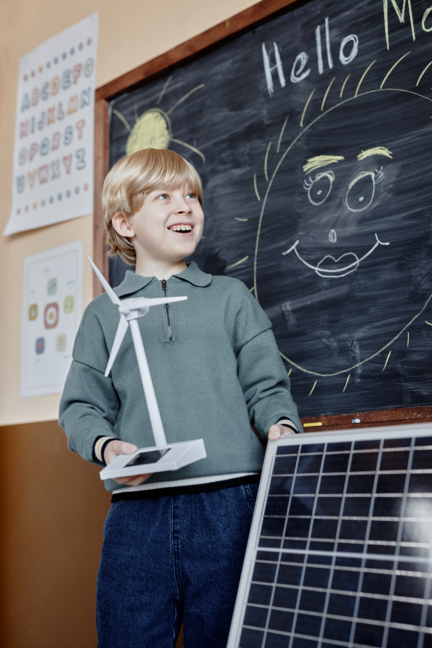 Vertical shot of cheerful primary learner presenting green energy project holding small solar powered wind turbine and big solar panel showing devices to class