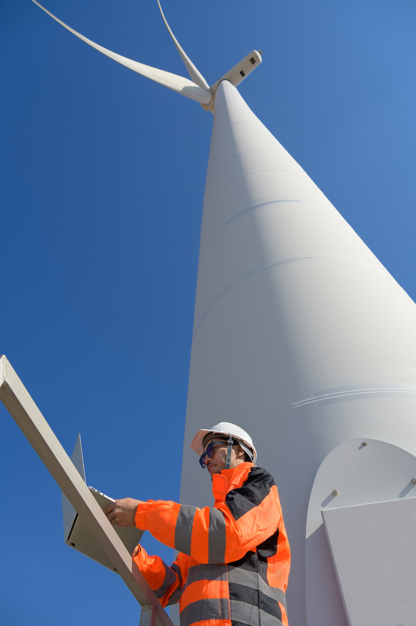 Male engineer in bright orange safety vest and white hard hat performing maintenance checks on white wind turbine by using laptop while standing against clear blue sky background. Renewable energy and clean technology concept